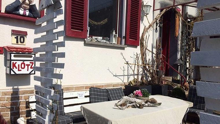 Een gezellige buitenruimte met een tafellaken en zitplaatsen. Op de achtergrond is er een vensterbank met planten en een decoratieve aap.