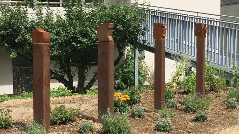 Un groupe de quatre colonnes rouillées se trouve dans un jardin. À l'arrière-plan, on peut voir des plantes et un bâtiment.