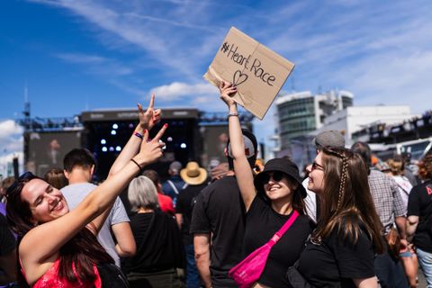 Eine fröhliche Menschenmenge auf einem Festival. Drei Frauen posieren mit einem Schild und genießen die Musik unter einem blauen Himmel.