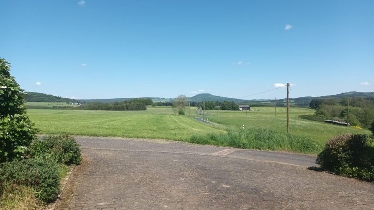 A peaceful landscape with green fields and gentle hills under a clear blue sky. A paved path leads into the expanses of nature.