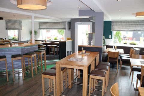 Ein modernes Restaurant mit Holztischen und Hochstühlen. Die Innenräume sind hell und einladend, mit großen Fenstern und einem gemütlichen Ambiente.