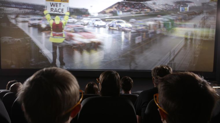 Ein Kinosaal mit Zuschauern, die ein Autorennen auf der Leinwand verfolgen. Ein Streckenposten hält ein Schild mit der Aufschrift "Wet Race".