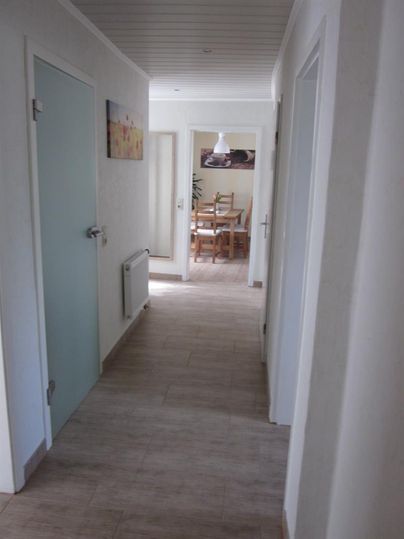 Een lichte gang met lichte muren en meerdere deuren. Aan het einde van de gang is een eetgedeelte zichtbaar met een tafel en stoelen.