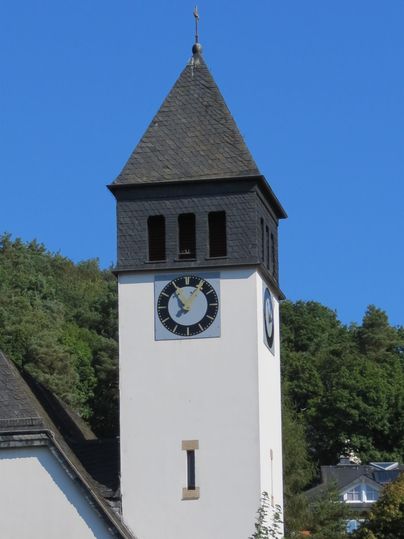 Ein Kirchturm mit einem schwarzen Dach und einer großen Uhr. Der Himmel ist klar und blau, umgeben von grünen Bäumen.