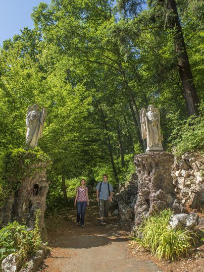 Ein schöner Weg im Wald, flankiert von beeindruckenden Statuen. Zwei Personen spazieren entspannt durch die grüne Umgebung.