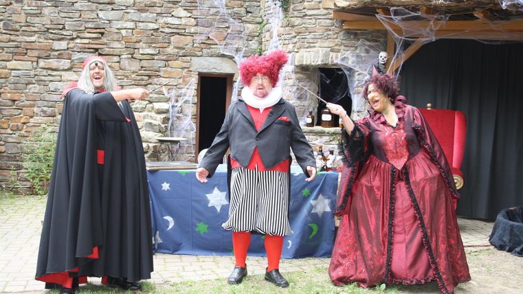 Drei Darsteller in fantasievollen Kostümen führen eine schrille Aufführung auf. Im Hintergrund sind Steine und eine Bühne zu sehen.