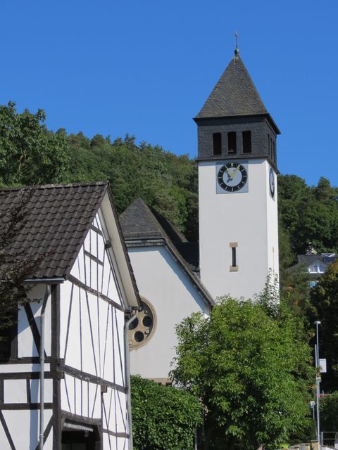 Eine Kirche mit einem hohen Turm und einer großen Uhr steht neben einem traditionellen Fachwerkhaus. Im Hintergrund sind grüne Hügel und ein blauer Himmel zu sehen.