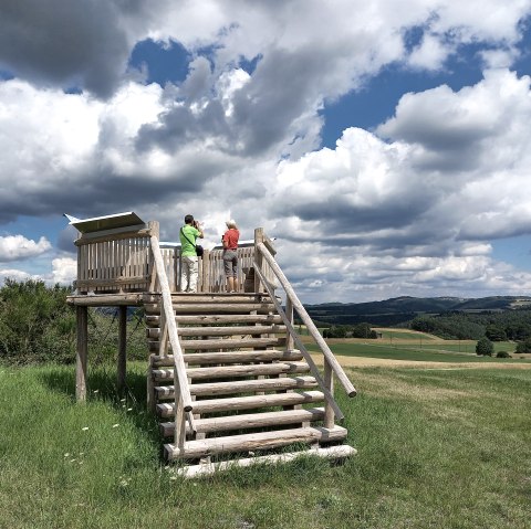 Deux personnes se tiennent sur une plateforme en bois et regardent le vaste paysage. Le ciel est en partie nuageux et en partie bleu., © Siegfried Müller