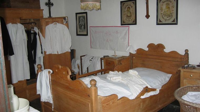 Ein einfaches Schlafzimmer mit einem Holzbett und weißen Bettdecken. Kleidung hängt in einem Schrank, und an der Wand sind Bilder und ein Kreuz zu sehen.
