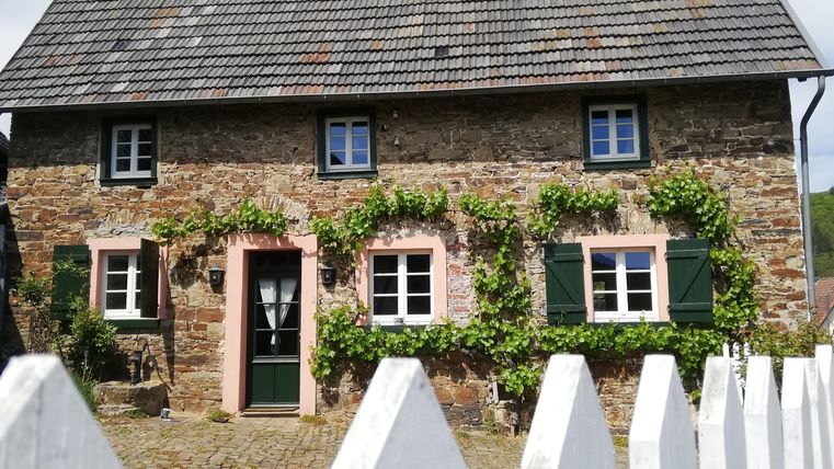 A charming stone house with pink window frames and green plants. The building has a slanted roof and is located behind a white fence.