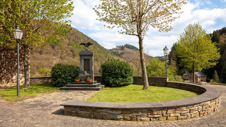 Ein gepflegter Platz mit einem Denkmal, umgeben von Bäumen und grünen Sträuchern. Im Hintergrund sind sanfte Hügel und ein blauer Himmel zu sehen.
