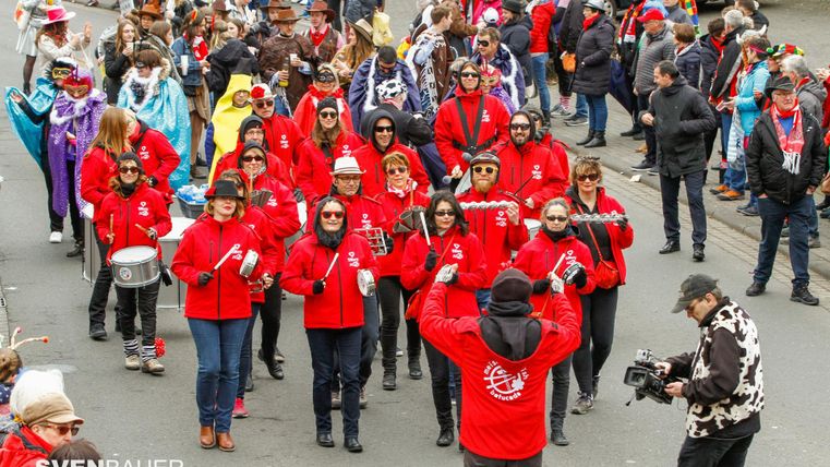 A colorful parade with many participants in red jackets. People are playing musical instruments and joyfully celebrating in the street.