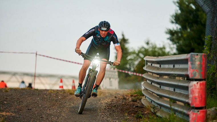 Ein Mountainbiker fährt auf einem schmalen Pfad und zeigt eine dynamische Fahrtechnik. Im Hintergrund sind Bäume und eine Rennstrecke zu sehen.