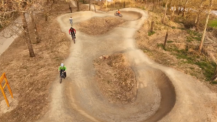 Pumptrack mit geschwungenen Wellen und Steilkurven, auf dem mehrere Personen mit Fahrrädern fahren, umgeben von Bäumen.