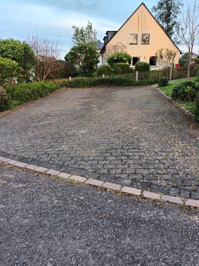 A paved driveway in front of a house with a garden. The surroundings are green and well-maintained.