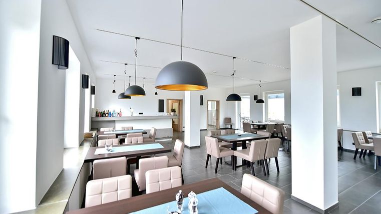 Ein modernes Restaurant mit hellen Wänden und eleganten Möbeln. Große Fenster sorgen für viel Tageslicht und eine einladende Atmosphäre.