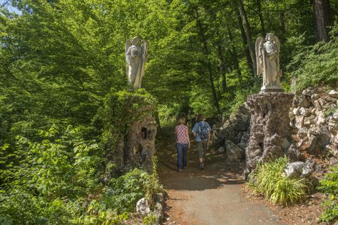 Ein malerischer Weg, gesäumt von grünen Bäumen und zwei steinernen Engel-Statuen. Zwei Personen schlendern gemütlich auf dem Pfad.