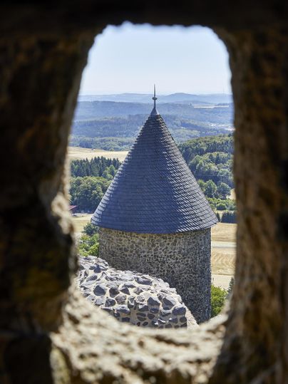 Ein Blick durch ein Steinfenster zeigt einen Turm mit einem spitzen Dach. Im Hintergrund sind sanfte Hügel und eine grüne Landschaft zu sehen.