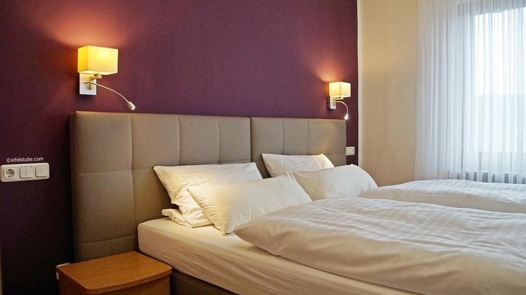 Une chambre d'hôtel confortable avec deux lits simples et des meubles modernes et simples. Les murs sont peints dans une teinte violette et il y a une grande baie vitrée avec des rideaux.