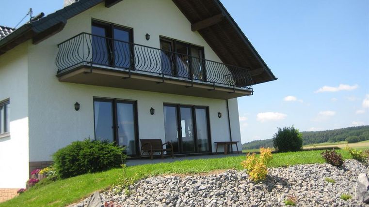 Une maison moderne avec un balcon et de grandes fenêtres. Le jardin bien entretenu avec des pierres et des plantes complète l'ensemble.