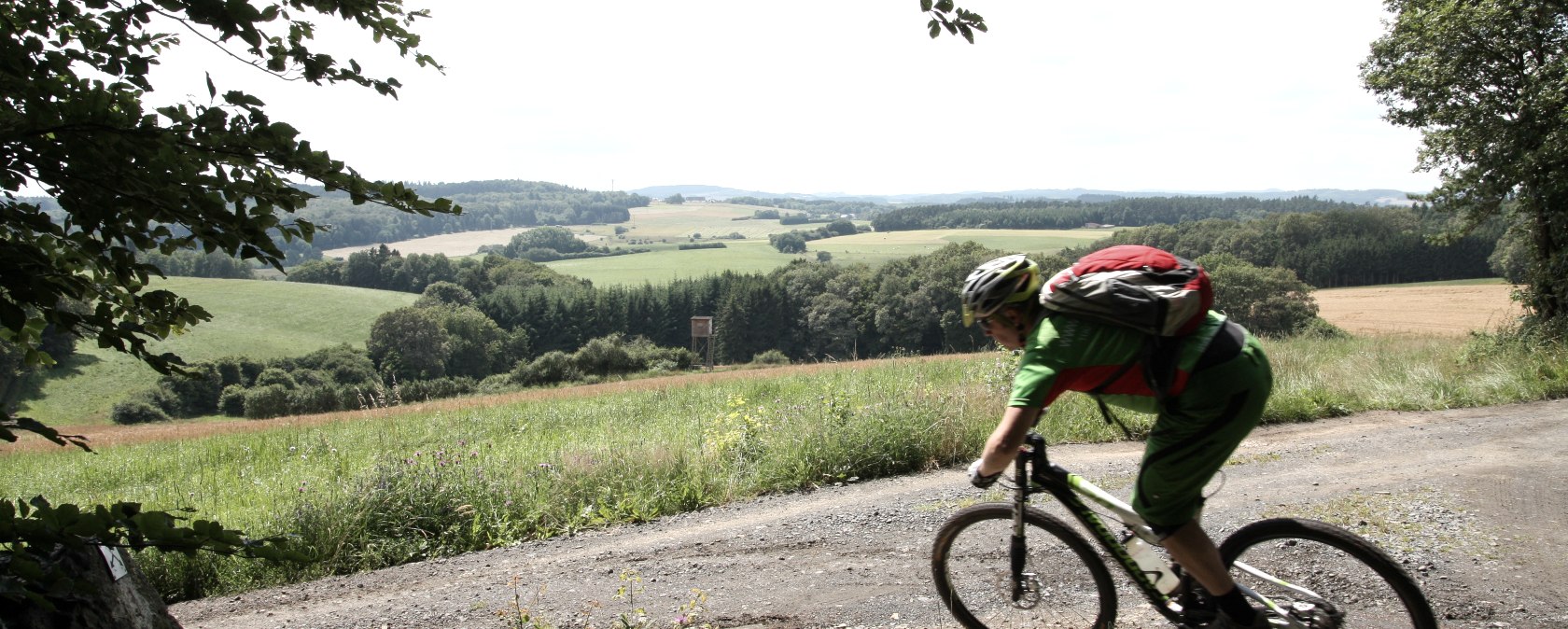 Ein Mountainbiker in gr&uuml;ner Kleidung f&auml;hrt auf einem Schotterweg durch eine gr&uuml;ne, h&uuml;gelige Landschaft. Im Vordergrund steht eine Bank., &copy; Schanze Communication