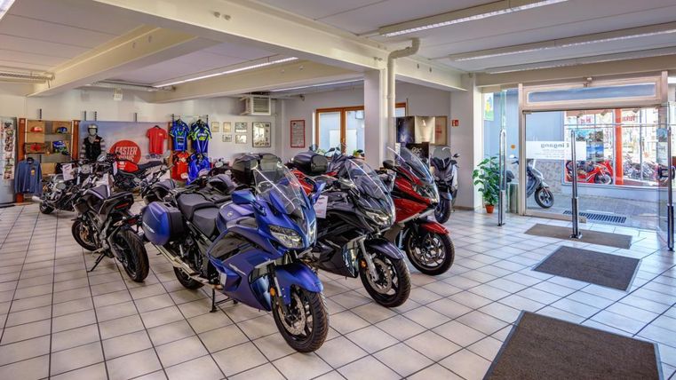 Ein Motorradgeschäft mit mehreren Motorrädern in verschiedenen Farben ausgestellt. Im Hintergrund sind Motorradbekleidung und Zubehör sichtbar.