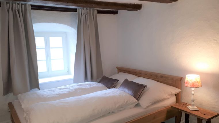 A cozy bedroom with a wooden bed and white bed covers. There is a window with curtains and a small lamp on a nightstand.