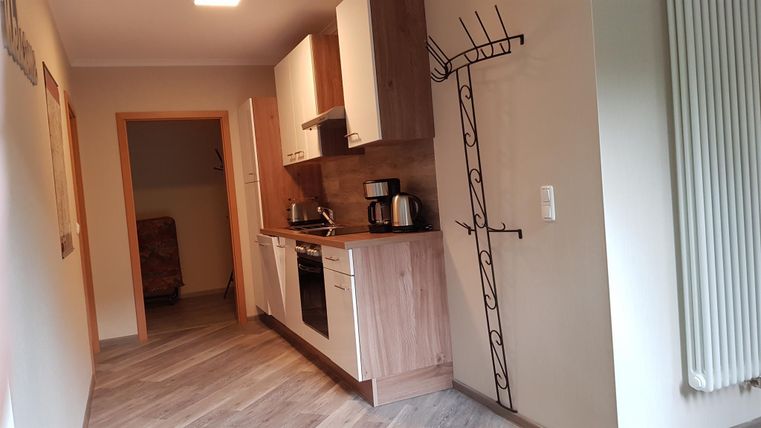 Een moderne keuken met houten kasten en lichte muren. Op de achtergrond is een gang met een kapstok te zien.