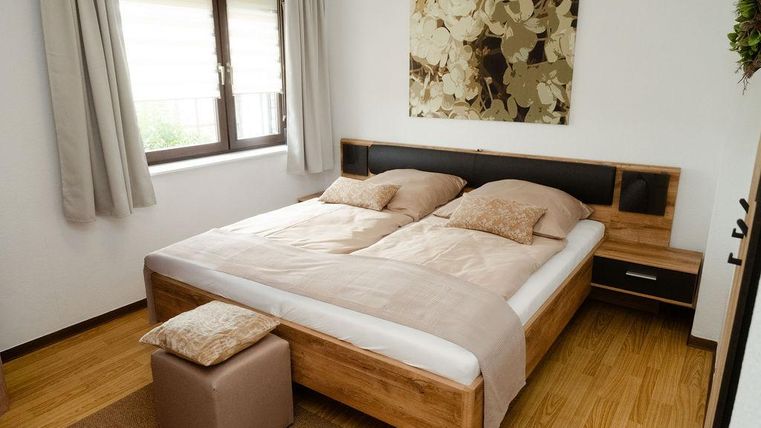 Ein modernes Schlafzimmer mit einem großen Bett und beigen Kissen. An der Wand hängt eine dekorative Wandkunst, während helle Vorhänge den Raum erhellen.