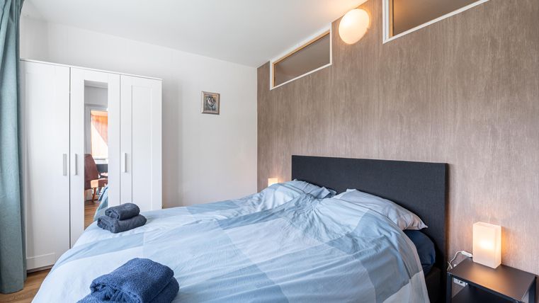 Een moderne slaapkamer met een groot bed en blauwe beddengoed. Een kast en nachtkastjes met lampen completeren het interieur.