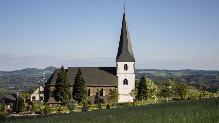 Une église avec un haut clocher se dresse dans un paysage pittoresque. Des prairies vertes et des collines douces entourent le bâtiment.