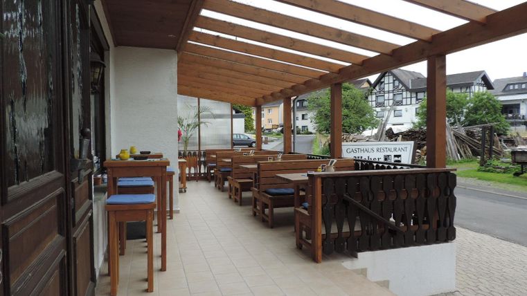 Eine einladende Terrasse mit Holztischen und Stühlen. Im Hintergrund sind Häuser und eine Grünfläche zu sehen.