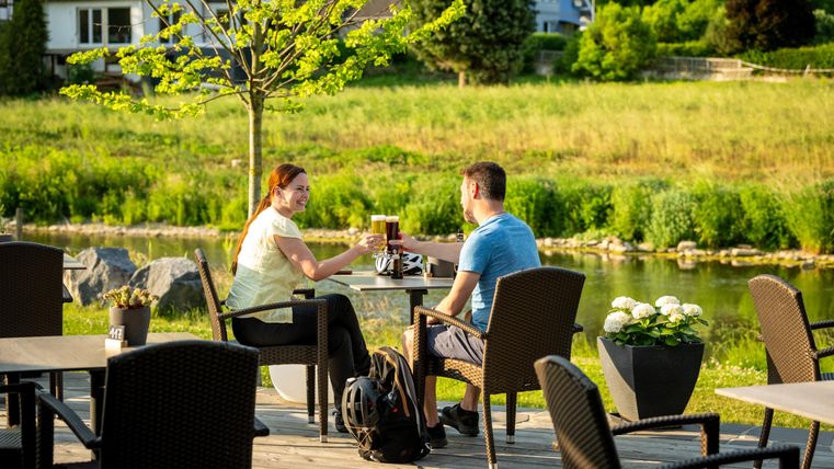 A couple is sitting at a table outdoors and clinking glasses. In the background, there are green meadows and trees visible.