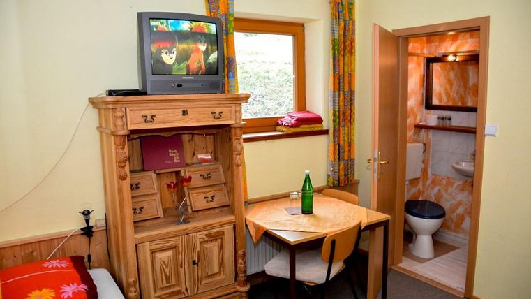 A simply furnished room with a television, a wooden wardrobe, and a small table. In the background, a bathroom is visible.