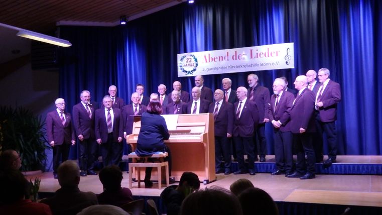 Ein Männerchor tritt auf einer Bühne auf, während ein Pianist sie begleitet. Im Hintergrund ist ein Banner mit der Aufschrift „Abend der Lieder“ zu sehen.