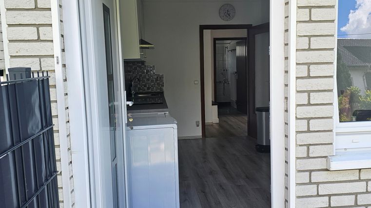 A view from the front door into a modern, bright room with wooden floor. Nearby, a kitchen with a refrigerator is visible.