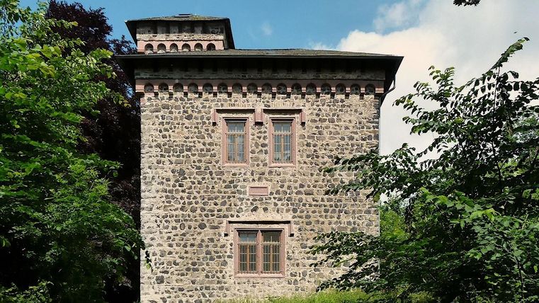 Ein historisches Steinhaus mit verzierten Fenstern, umgeben von üppigem Grün. Der Himmel ist klar mit wenigen Wolken.