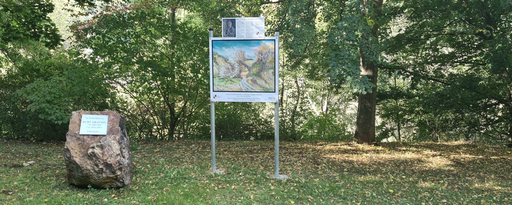 Ein Gedenkplatz im Wald mit einem Gedenkstein f&uuml;r Kurt Gratzel und einem Bild auf einer Tafel. Umgeben von B&auml;umen und gr&uuml;nem Laub., &copy; Tourist-Information Hocheifel-N&uuml;rburgring