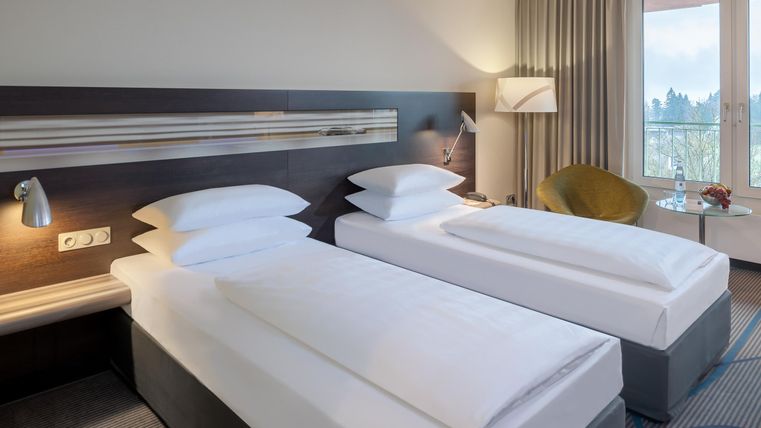 A modern hotel room with two single beds and elegant decor. The room features a large window with a view of the surroundings.