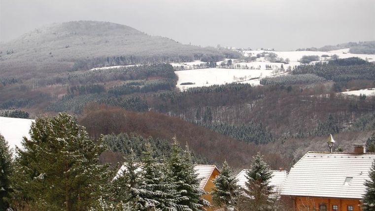 Een besneeuwd landschap met zachte heuvels en groene naaldbomen. Op de voorgrond zijn enkele houten gebouwen te zien, omringd door sneeuw.