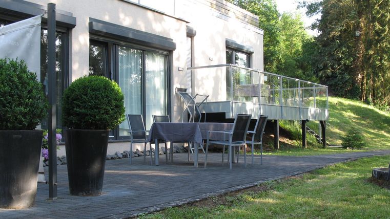 Ein modernes Haus mit einer Terrasse und Gartenmöbeln. Die Umgebung ist grün und gepflegt.