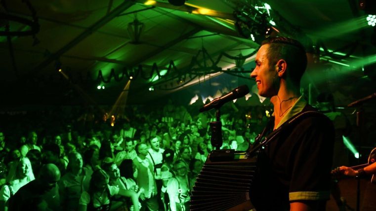 A musician is playing on stage in front of an enthusiastic crowd. The atmosphere is lively and illuminated with green lights.