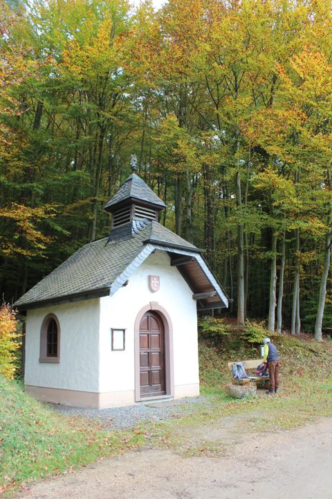 Eine kleine Kapelle steht im Wald mit bunten Bäumen im Hintergrund. Neben der Kapelle ist eine Person mit einem Stuhl beschäftigt.