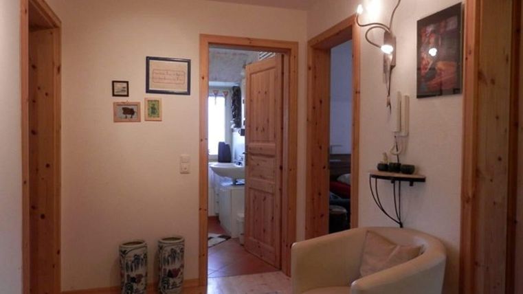 Een lichte gang met houten details en meerdere deuren. Aan de rechterkant staat een comfortabele stoel en er hangt een wandlamp.