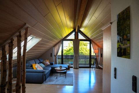 Ein gemütliches Wohnzimmer in einem Dachgeschoss mit Holzdecke. Große Fenster bieten einen Blick auf den Außenbereich und lassen viel Tageslicht herein.