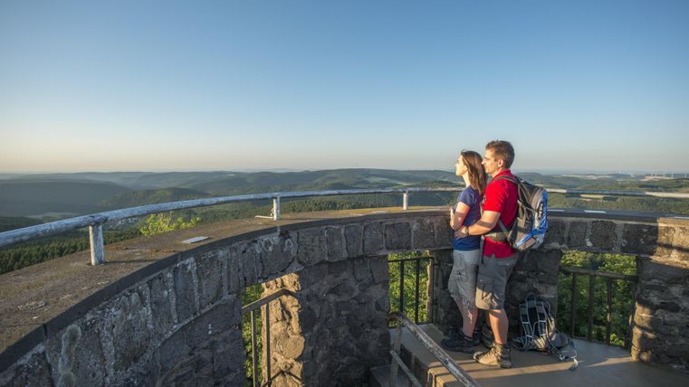 Ein Paar steht auf einer Aussichtsplattform und schaut in die Ferne. Die Landschaft ist hügelig und von Bäumen umgeben, während der Himmel klar und blau ist.