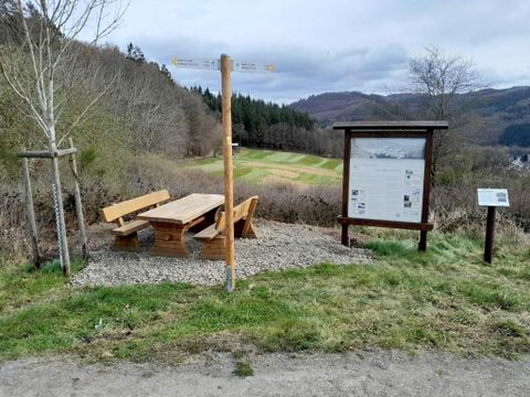 Une zone de pique-nique avec un banc en bois et une table dans un cadre rural. À l'arrière-plan, on peut voir des arbres et une prairie.