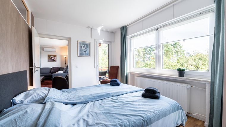 Een lichte slaapkamer met een tweepersoonsbed en grote ramen. De inrichting is modern en vriendelijk met uitzicht op de buitenlucht.