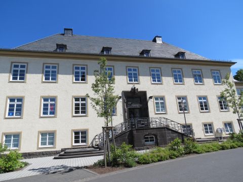 Ein historisches Gebäude mit einem geräumigen Eingangsbereich und großen Fenstern. Die Fassade ist hell und von Bäumen umgeben.