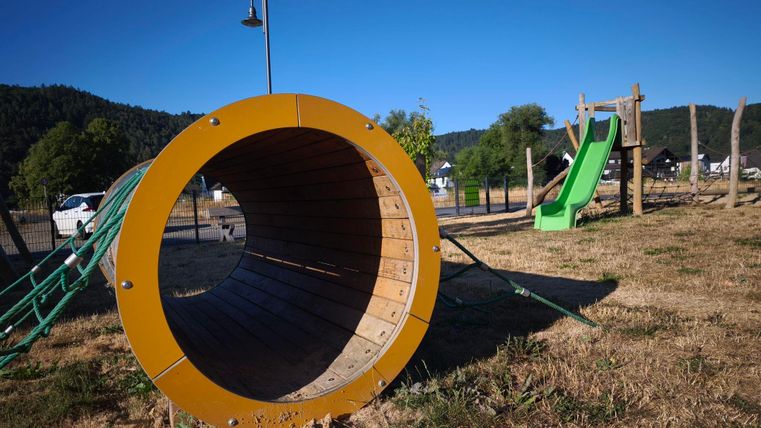 Ein Spielplatz mit einer großen, runden Röhre und einer grünen Rutsche. Im Hintergrund sind Bäume und eine klare blaue Himmel zu sehen.